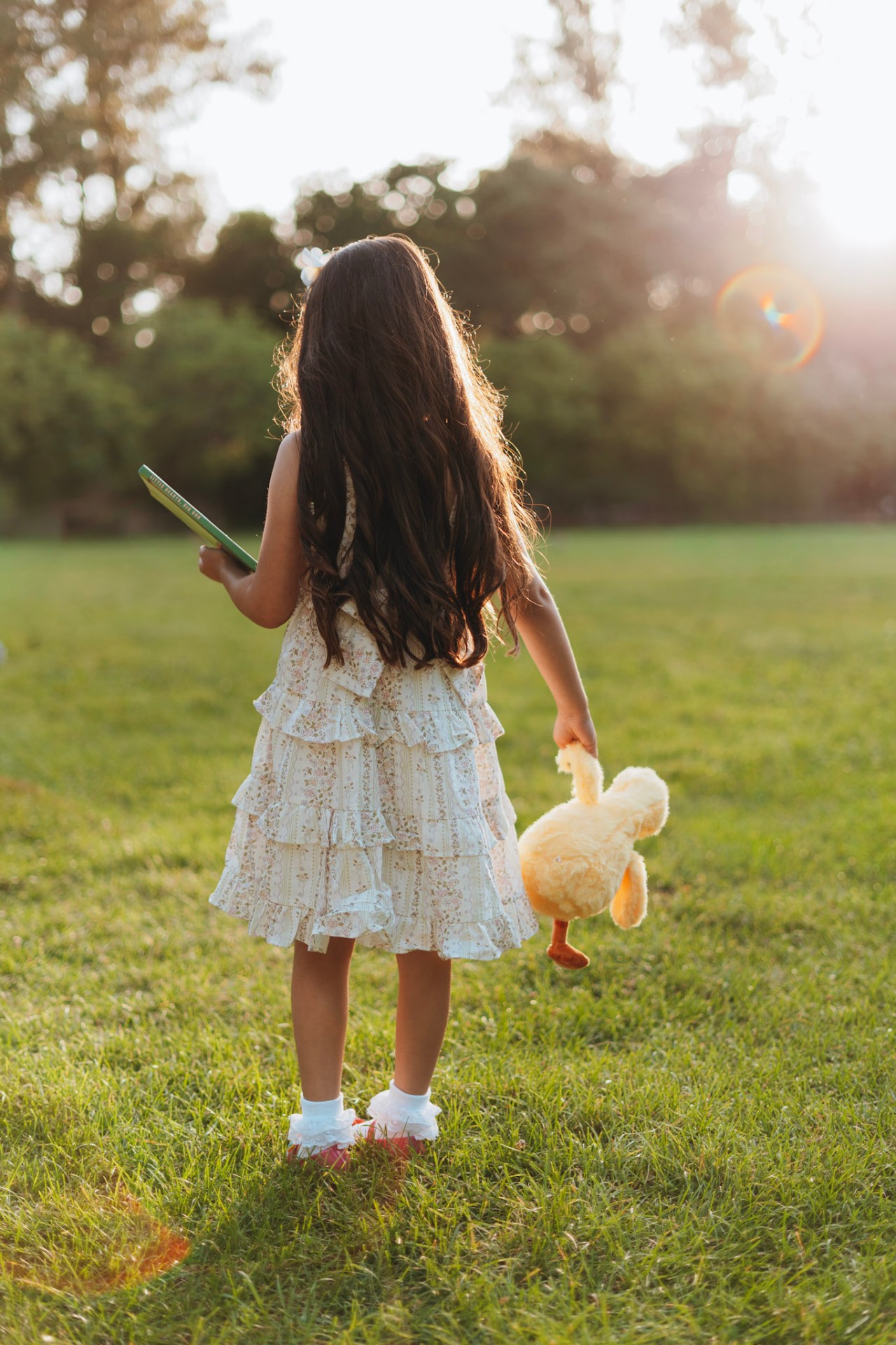 A little girl in a floral dress holding Little Hearts, Big God and a plush duck in a sunlit field